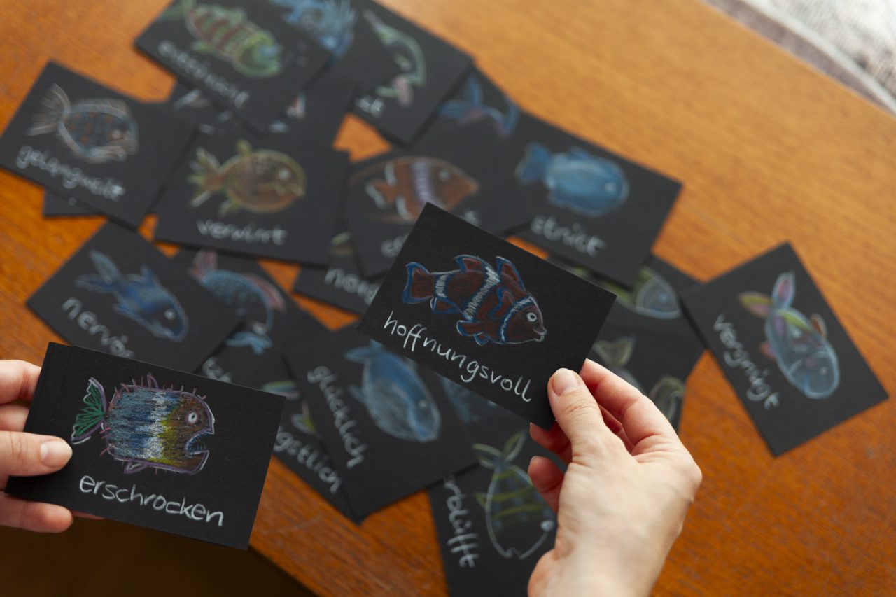 karteikarten Hände halten zwei schwarze Karten mit Fischzeichnungen und den Worten "hoffnungsvoll" und "erschrocken", weitere Karten liegen verstreut auf einem Holztisch.